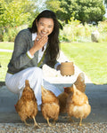 A woman crouches outdoors on a sandstone path, smiling and holding a small pottery container and feeding four brown chickens which are standing in front of her in a sunny garden setting.