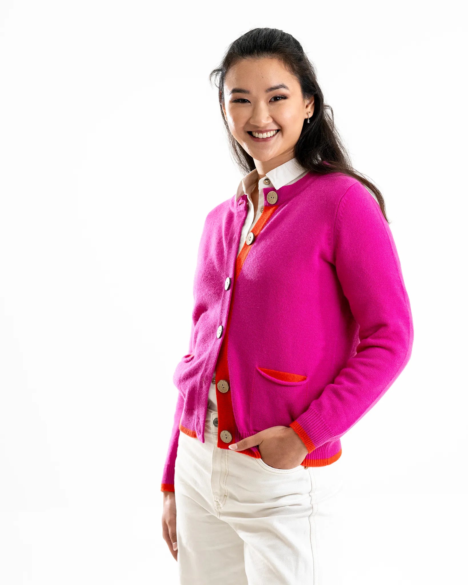 Woman wearing a bright pink cashmere cardigan over a white shirt with orange trim on a white background