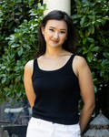 Woman wearing a pure cashmere black camisole tank top vest and white trousers standing against a pillar outdoors with greenery in the background