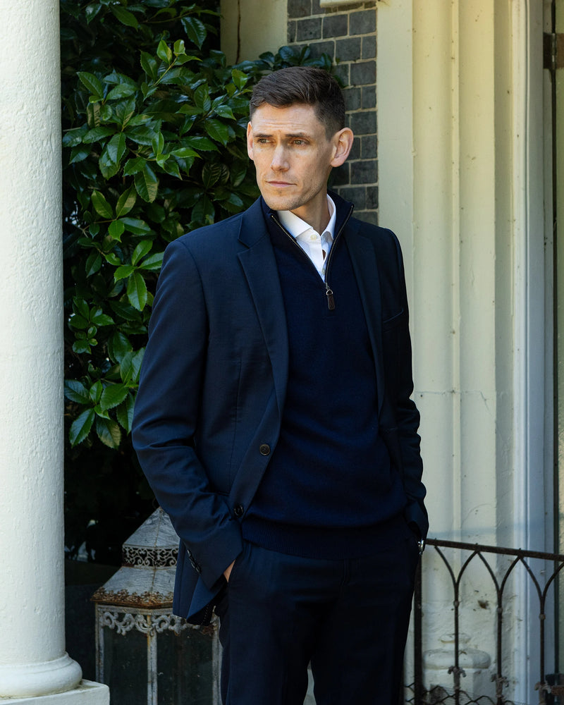 A man wearing a dark navy blue blazer, matching cashmere sweater, and trousers over a white collared shirt, standing outdoors in front of a white column and green foliage.