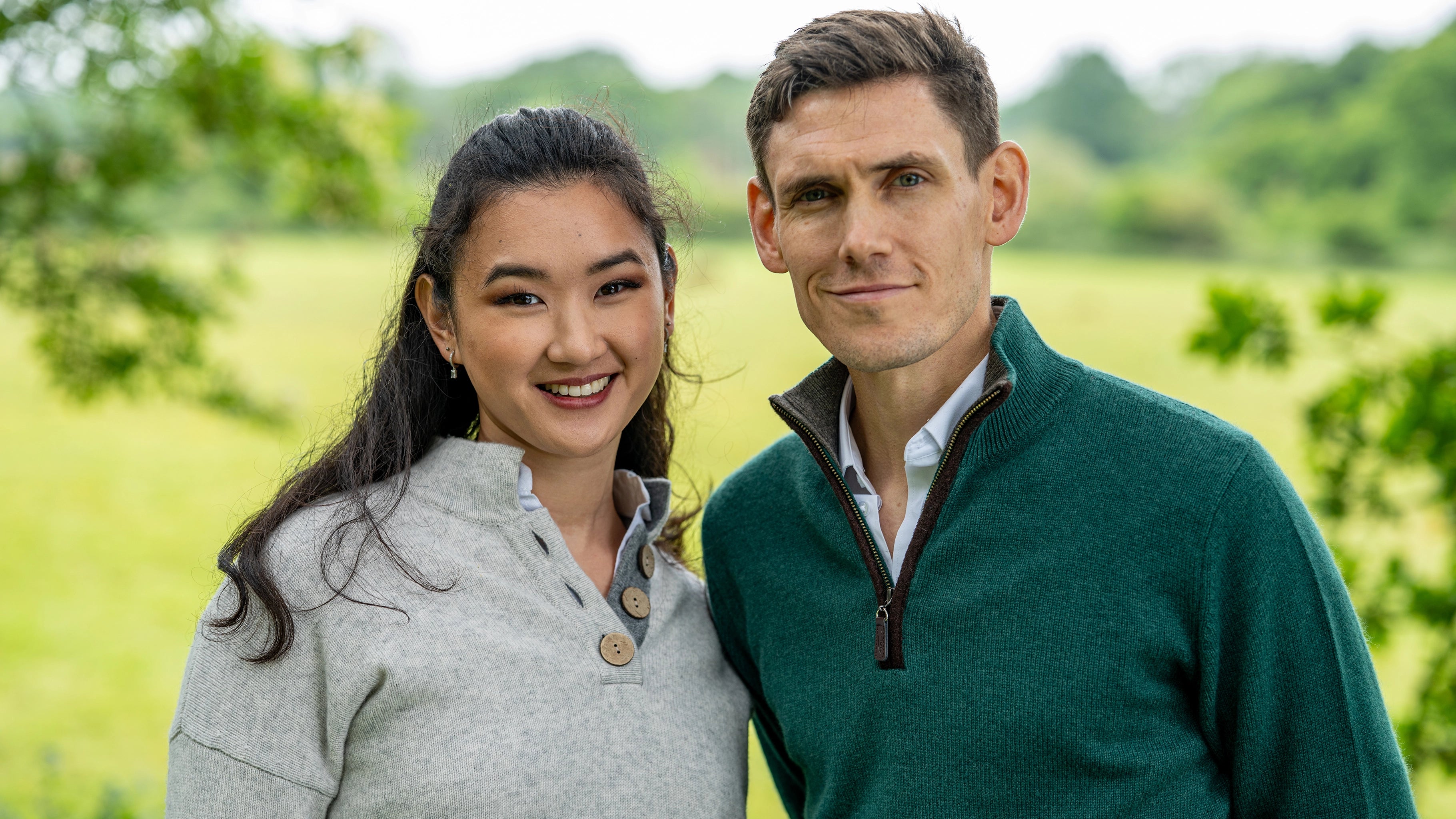 Two people standing together in a green outdoor setting with woman wearing a grey cashmere sweater and the man wearing a green cashmere sweater