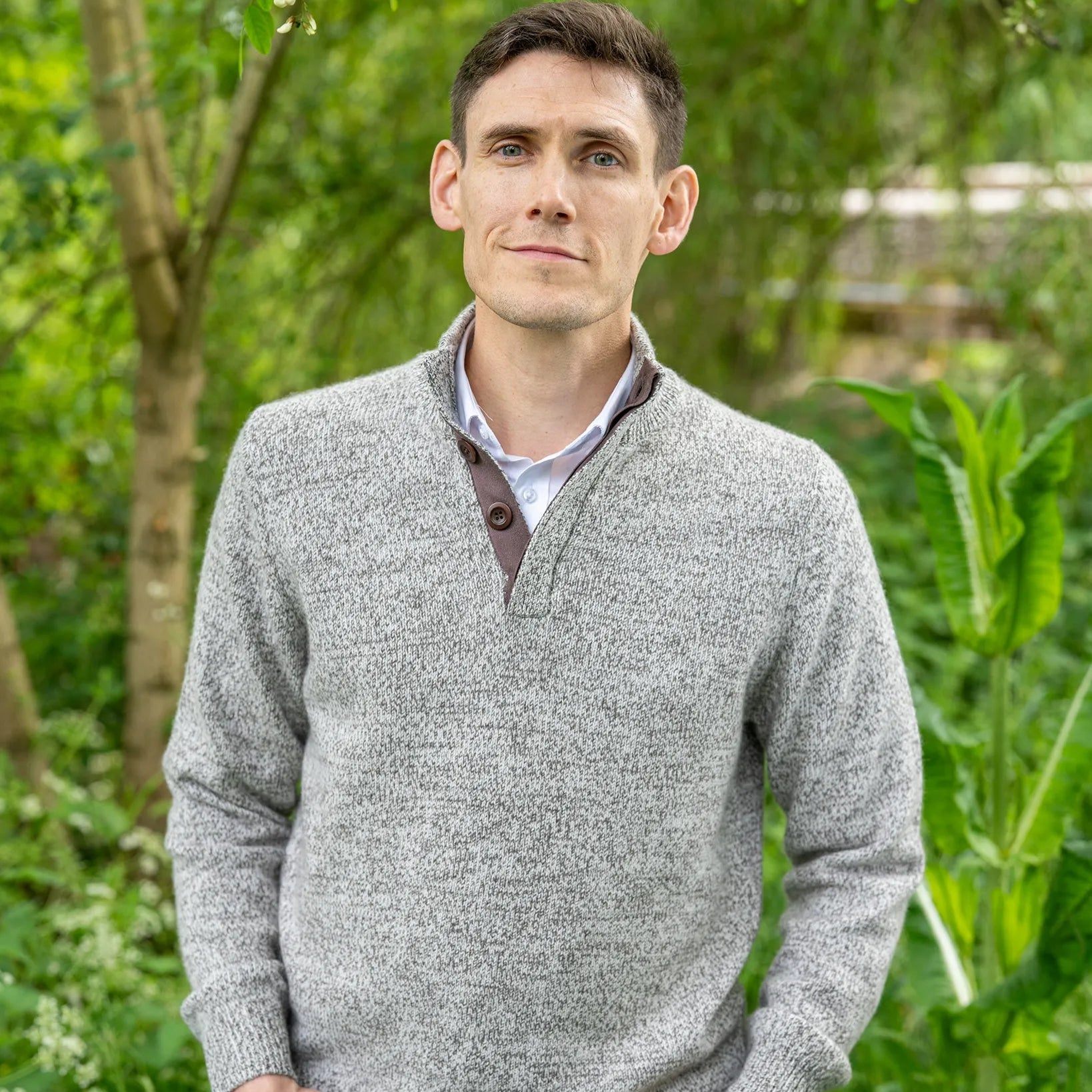 Man wearing a grey sweater in cashmere and merino marl and brown corozo buttons standing in a garden