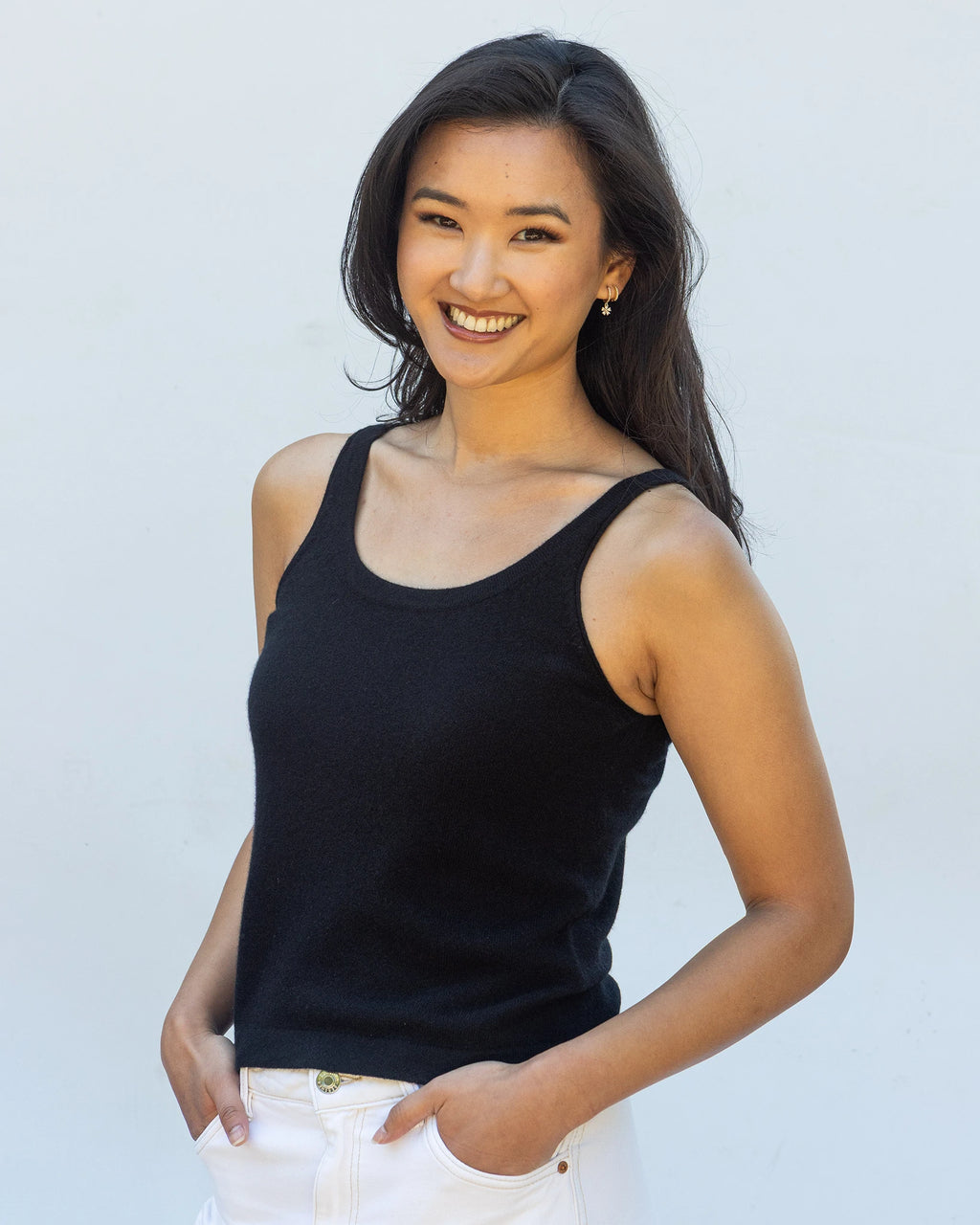 A smiling woman with long dark hair wearing a pure cashmere tank top in black with white trousers, standing against a plain light grey background.