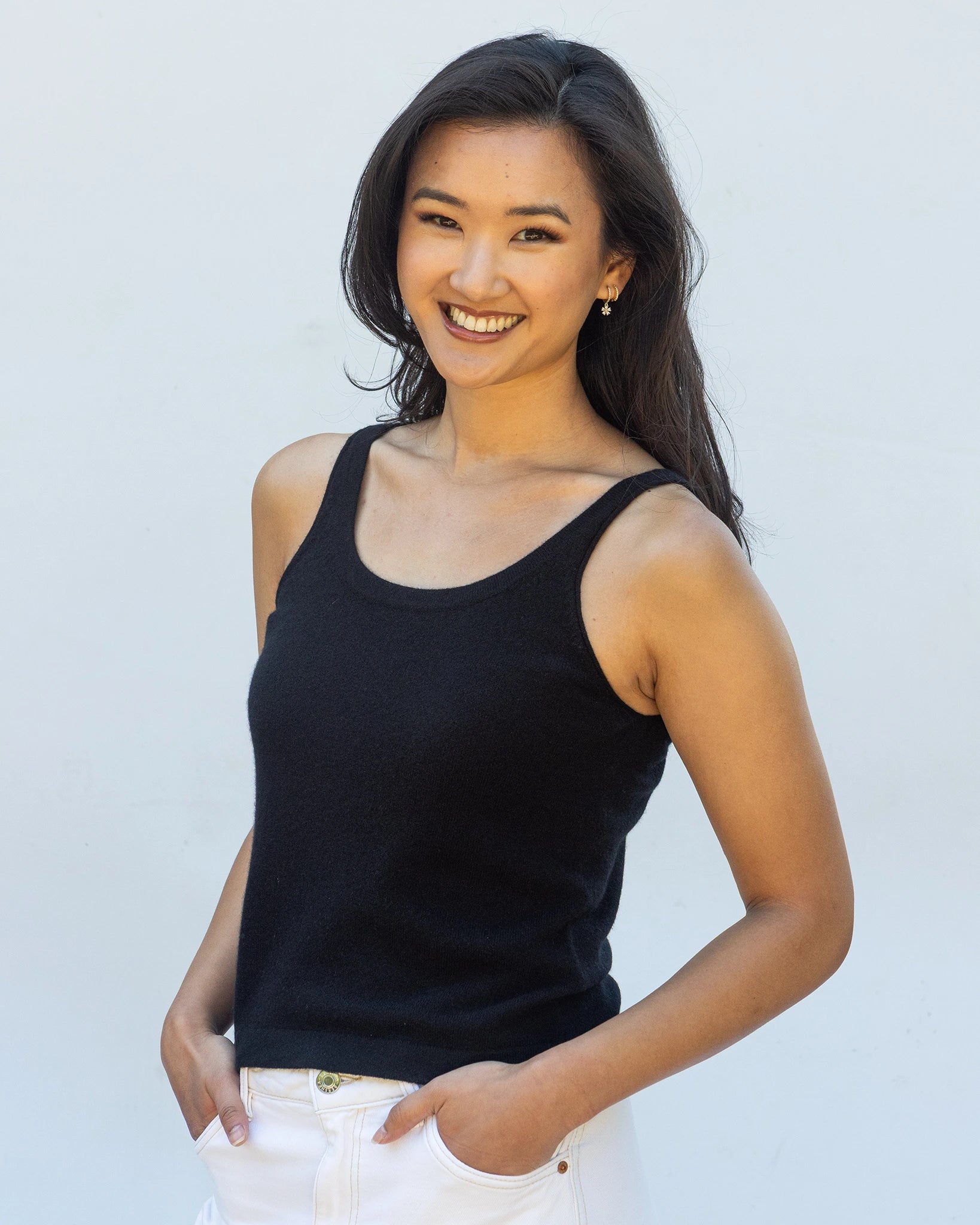 A smiling woman with long dark hair wearing a pure cashmere tank top in black with white trousers, standing against a plain light grey background.