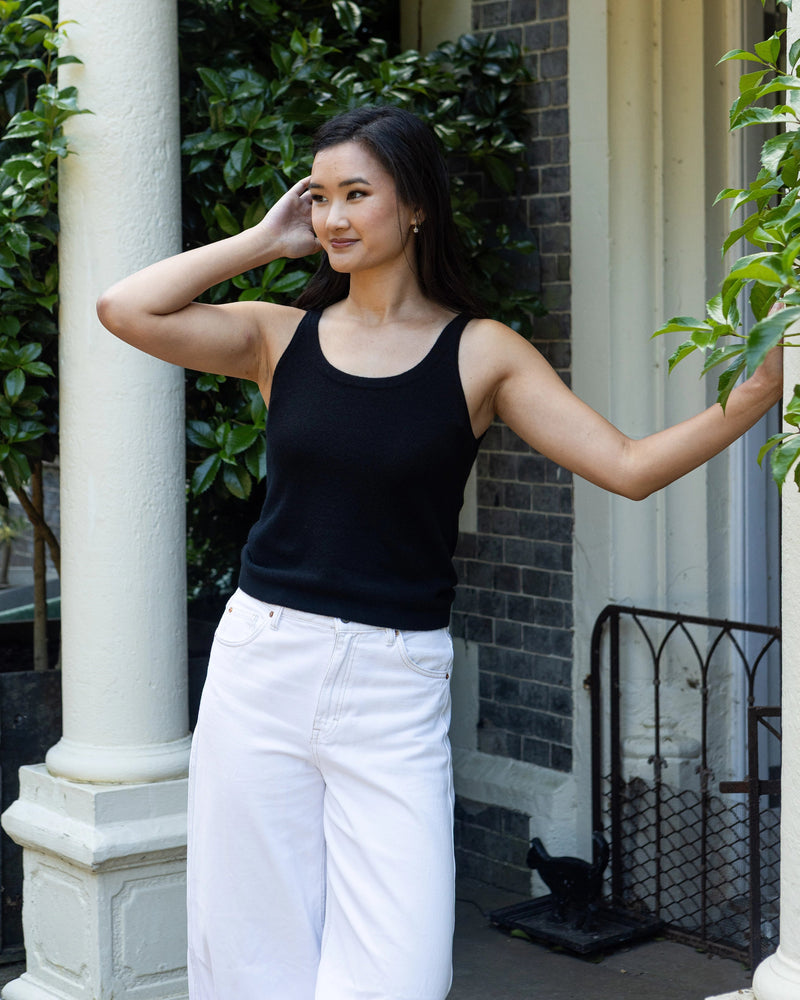 A woman wearing a black pure cashmere vest camisole tank standing in front of a building with white columns, brick walls, and green plants.