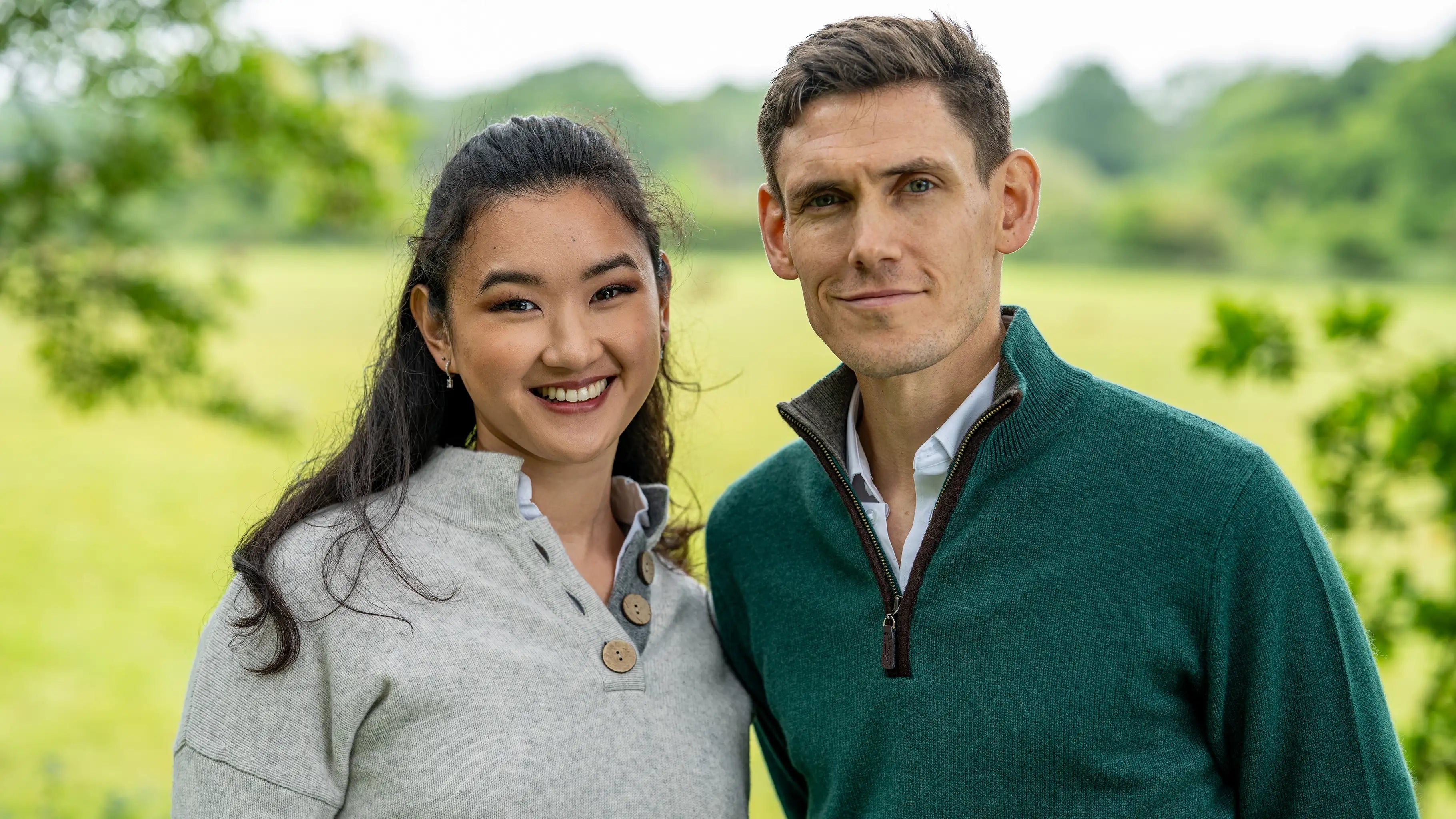 Two people standing together in a green outdoor setting with woman wearing a grey cashmere sweater and the man wearing a green zip neck sweater