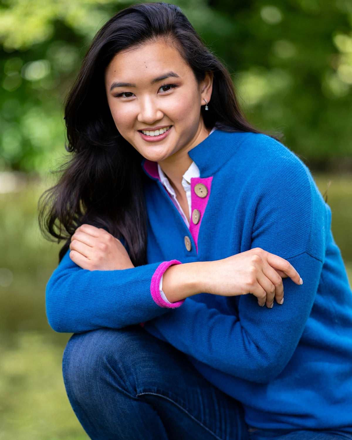 Model wearing high collar jumper in Aztec blue with pink collar and cuffs and large coconut buttons by river setting