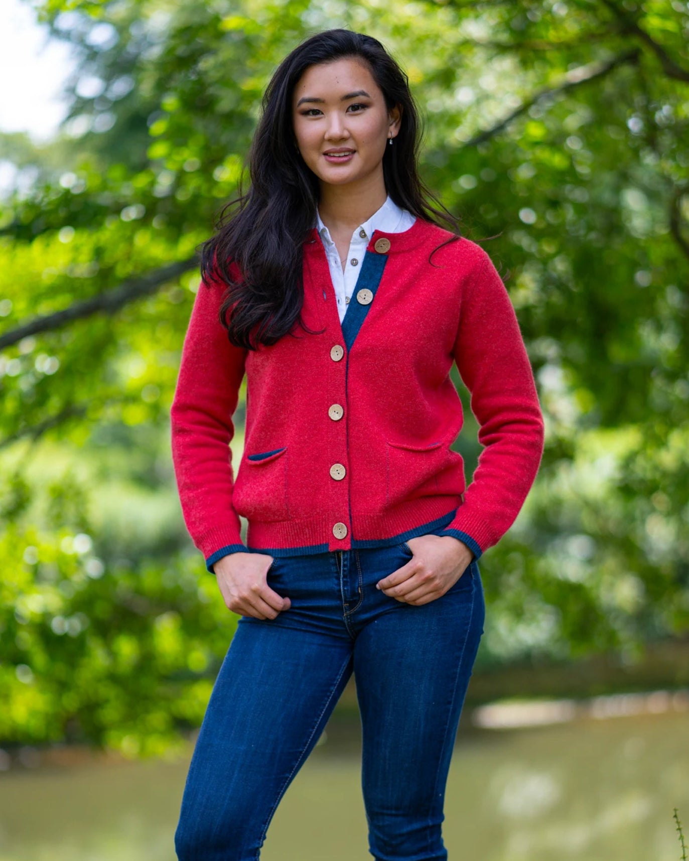 A woman standing outdoors wearing a burnt coral pocket cardigan with large coconut buttons, paired with blue jeans.