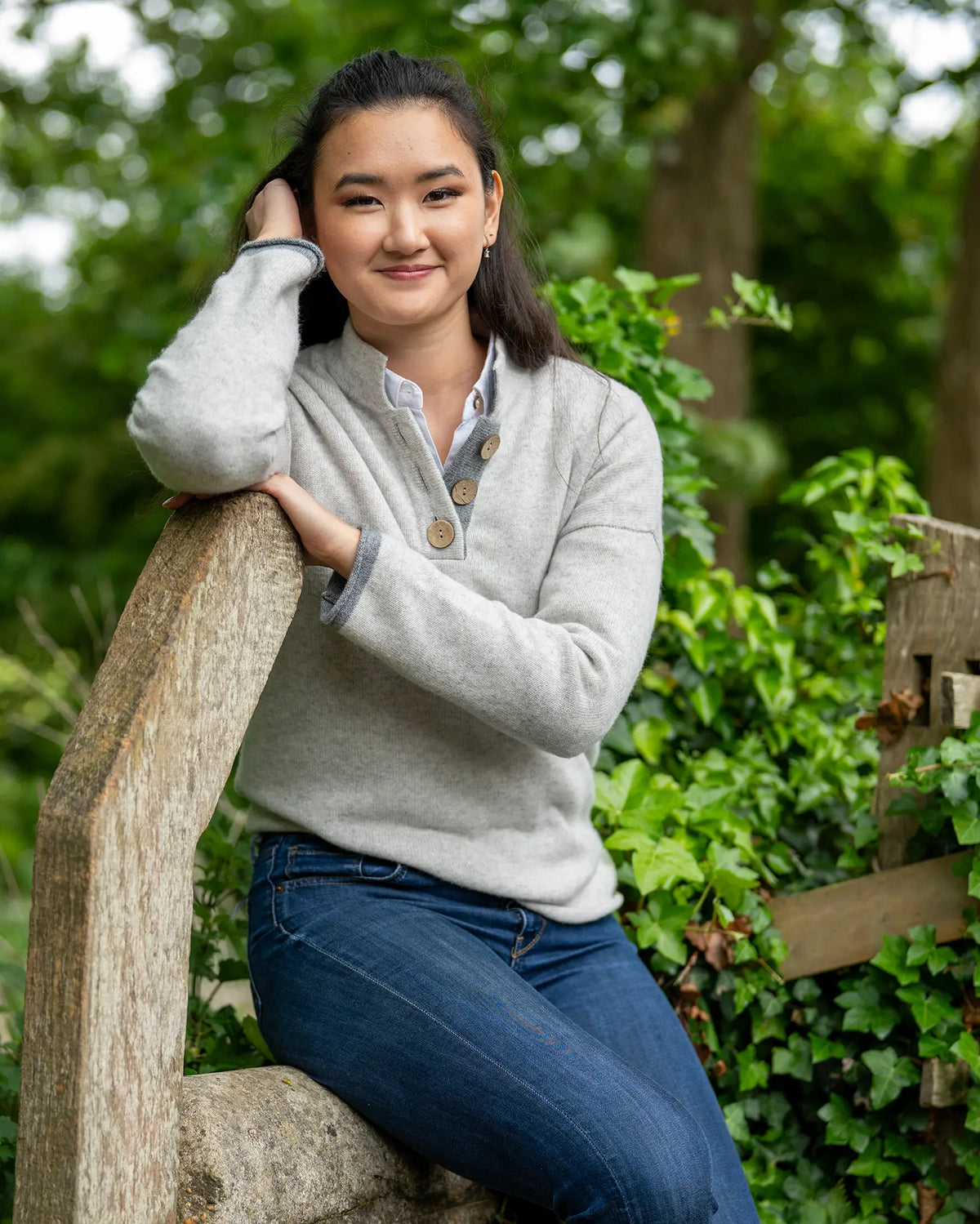 Woman in a cashmere grey sweater and blue jeans sitting on a wooden bench with greenery in the background