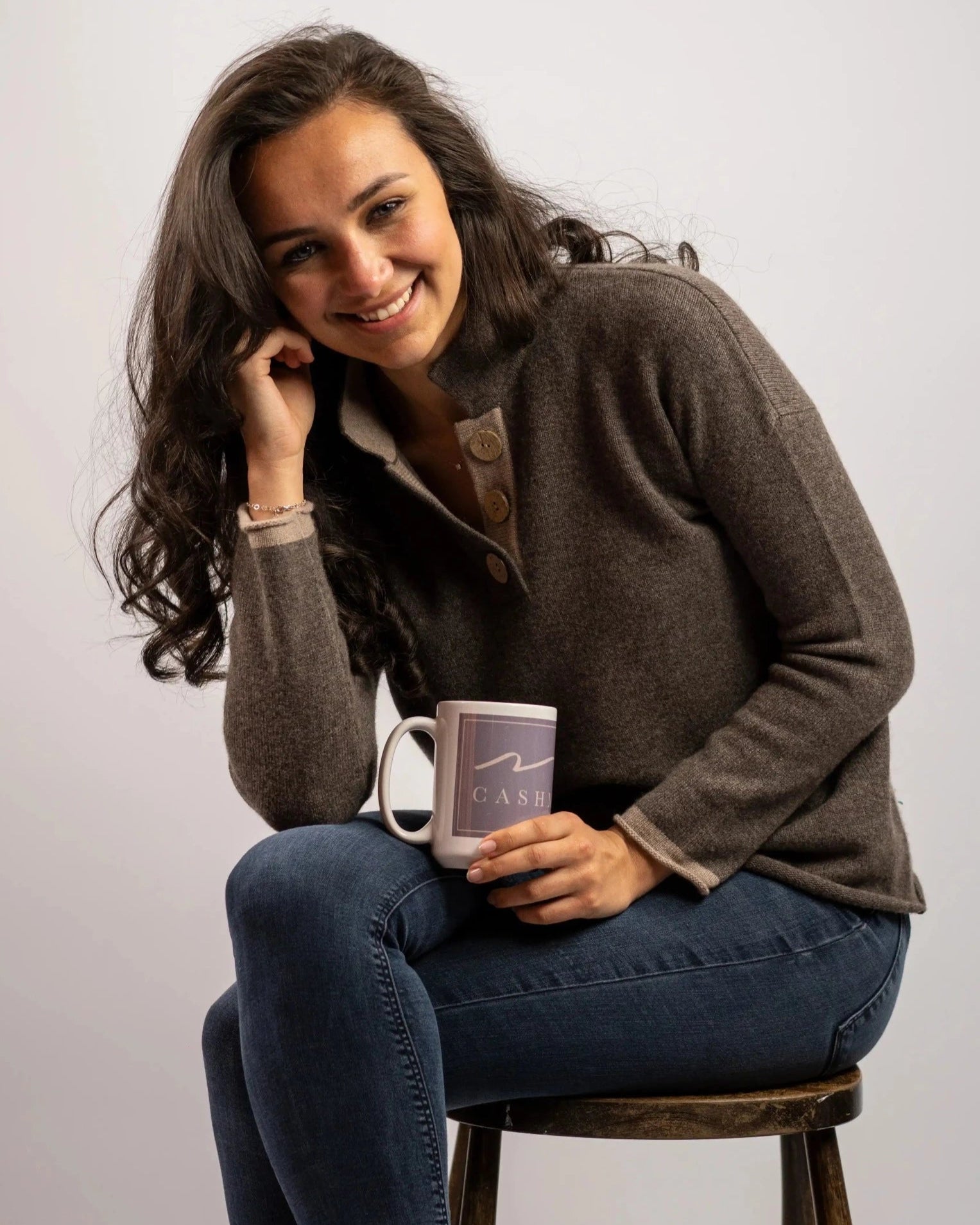 Model sitting on stool wearing cashmere and merino jumper in mole with large coconut buttons