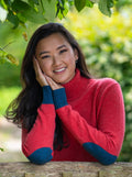 A woman smiling and wearing a burnt coral roll neck jumper with blue elbow patches and cuffs, standing outdoors.