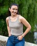 Woman wearing a beige cashmere vest and blue jeans standing outdoors with greenery in the background