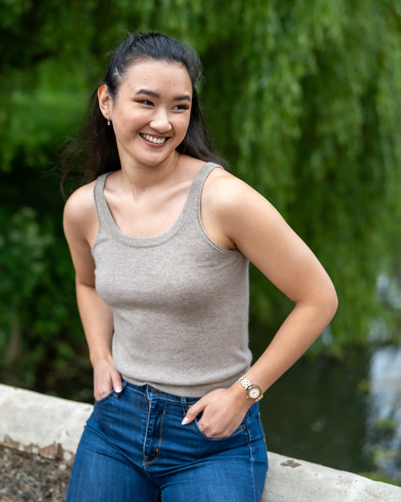 Woman wearing a beige cashmere vest and blue jeans standing outdoors with greenery in the background