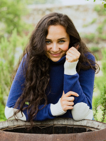 Women's roll neck jumper in blue with cream elbow patches, cuffs and bottom hem in cashmere merino leaning on a water tub by Misty Cashmere.